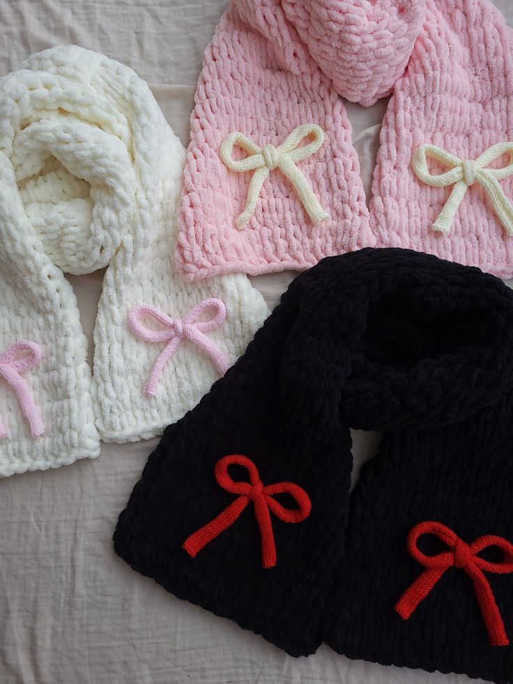 three knitted hats and scarfs are laying on a white sheet with red bows