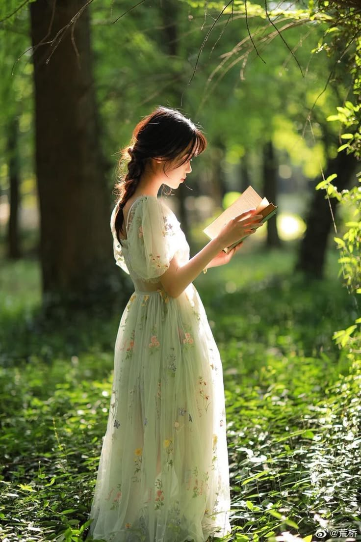 a woman in a green dress reading a book while standing in the grass with trees behind her