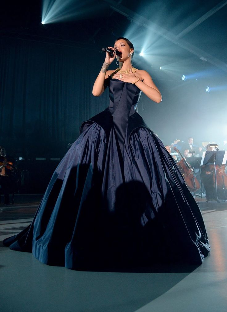 a woman in a black dress on stage with microphone and orchestra behind her, performing