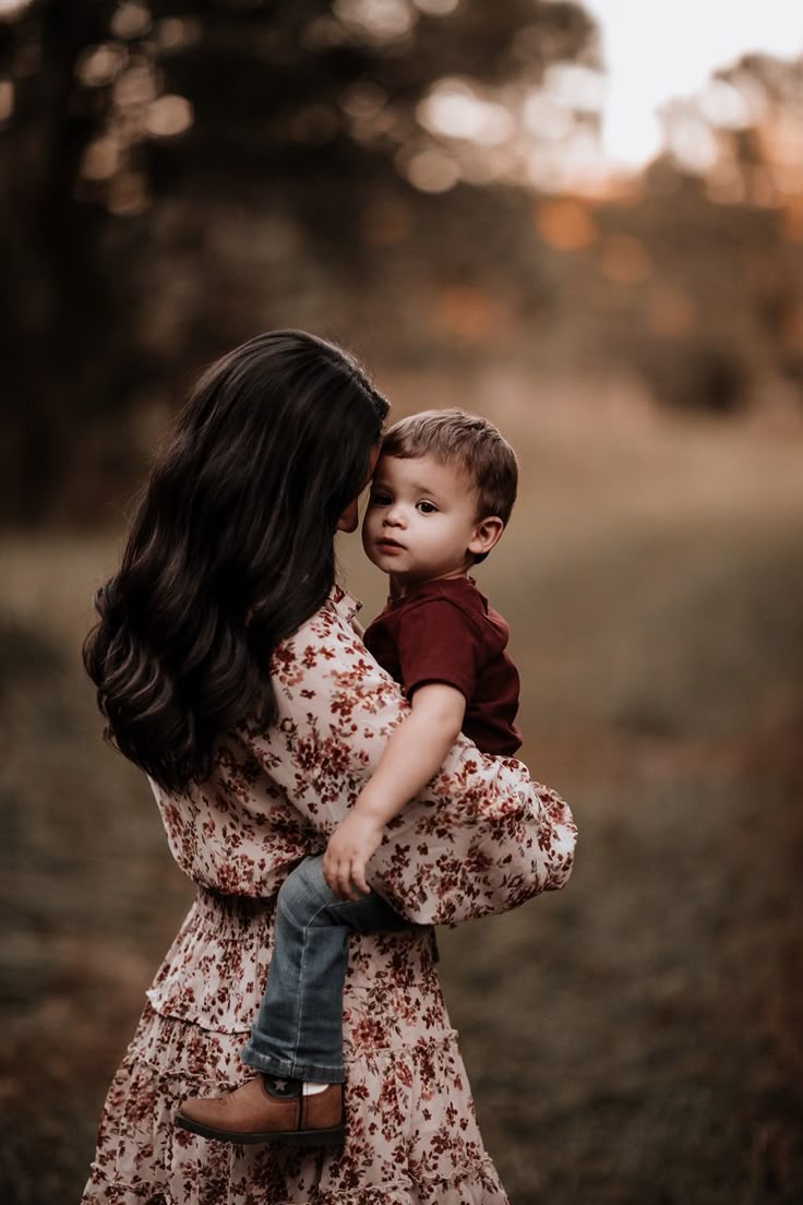 a woman holding a small child in her arms while standing in a field with trees