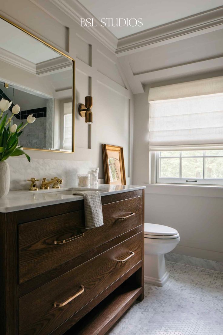 a white toilet sitting next to a wooden dresser in a bathroom under a window with roman shades