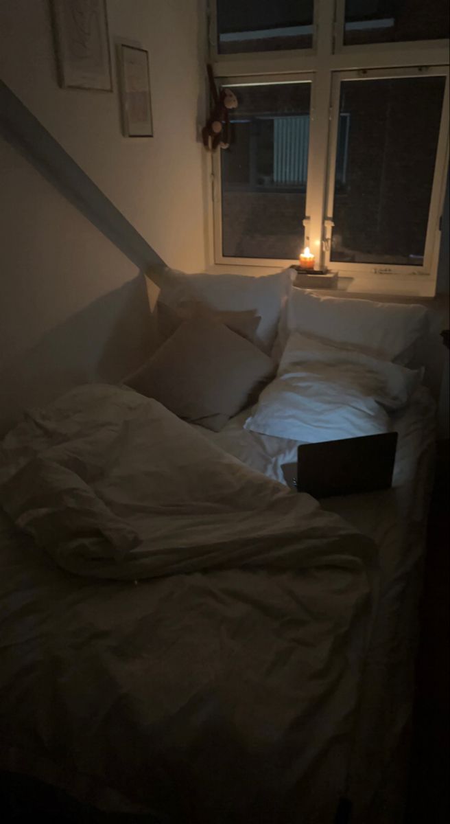 an unmade bed with white sheets and pillows in front of a window at night