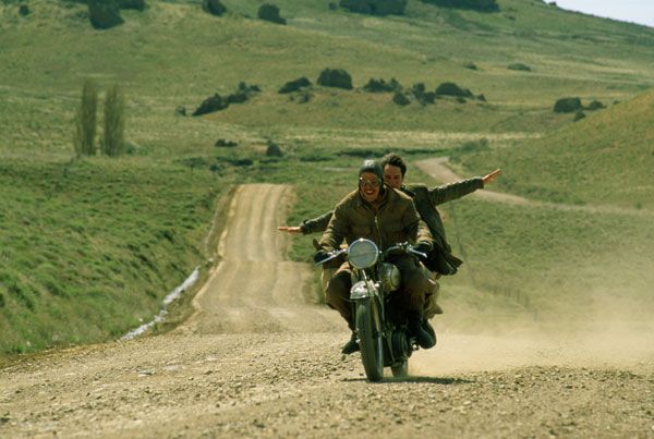 two men riding on the back of a motorcycle down a dirt road