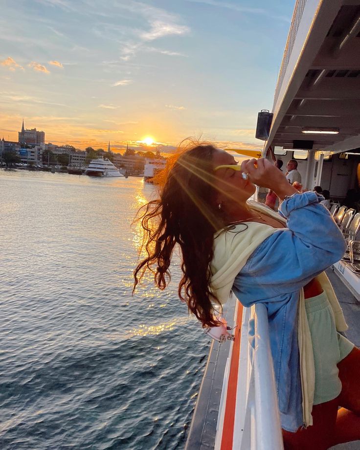 a woman standing on the side of a boat looking at the sun setting behind her