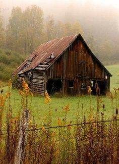 an old barn sits in the middle of a foggy field with tall grass and trees