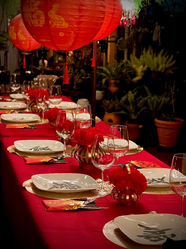 a long table with plates and wine glasses on it, lit by red lanterns in the background