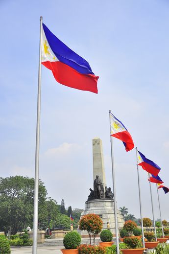 several flags flying in the wind next to a statue and flowerbeds with trees