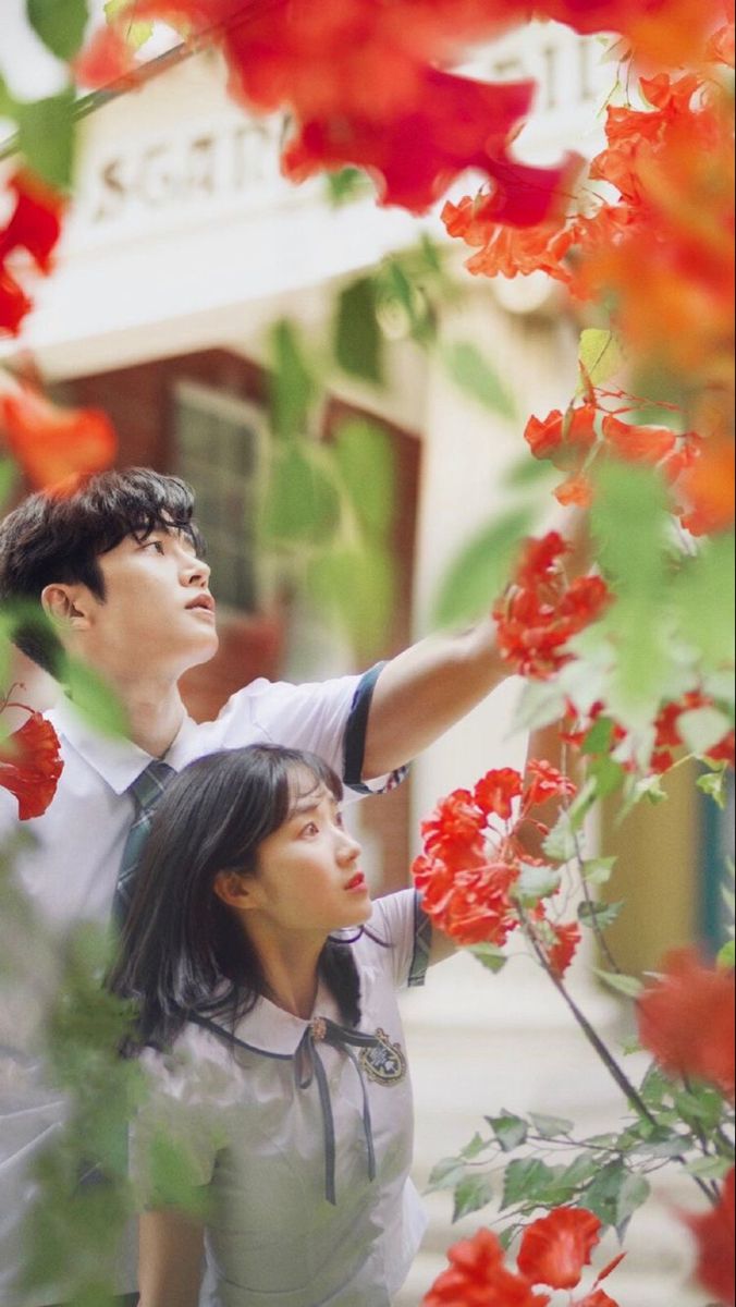 a man and woman standing next to each other in front of red flowers on a tree