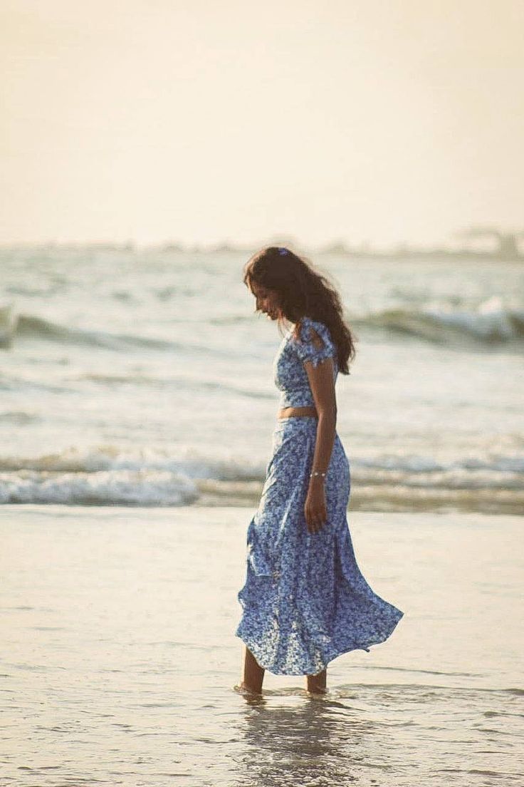 a woman in a blue dress standing on the beach looking down at the water's edge