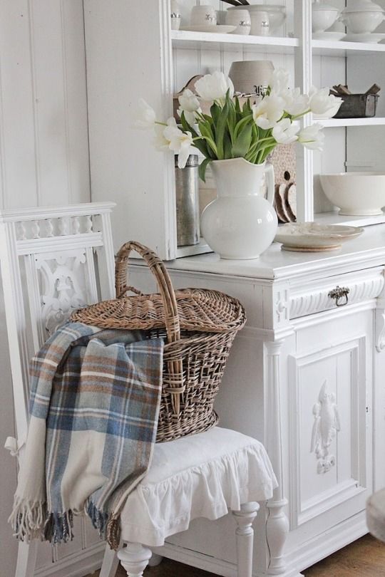 a white table topped with a basket filled with flowers next to a chair and mirror