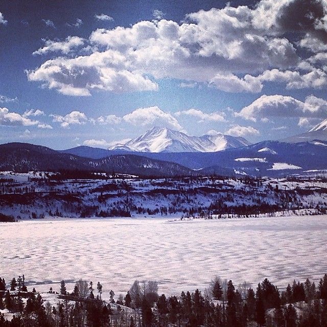 the mountains are covered in snow and trees under a blue sky with fluffy white clouds