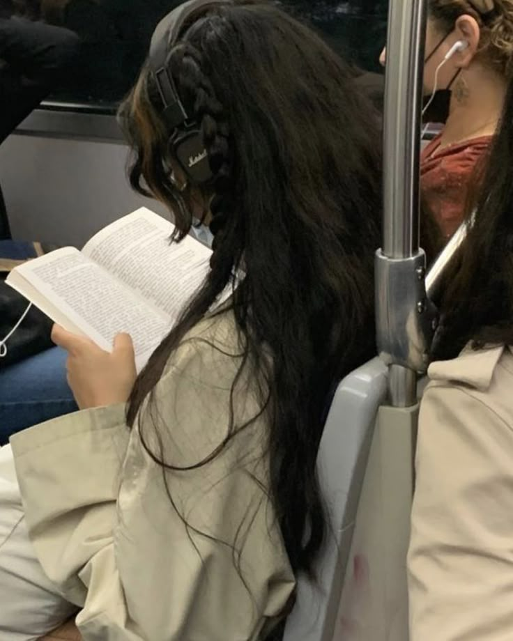 a woman with long hair sitting on a bus reading a book and listening to headphones