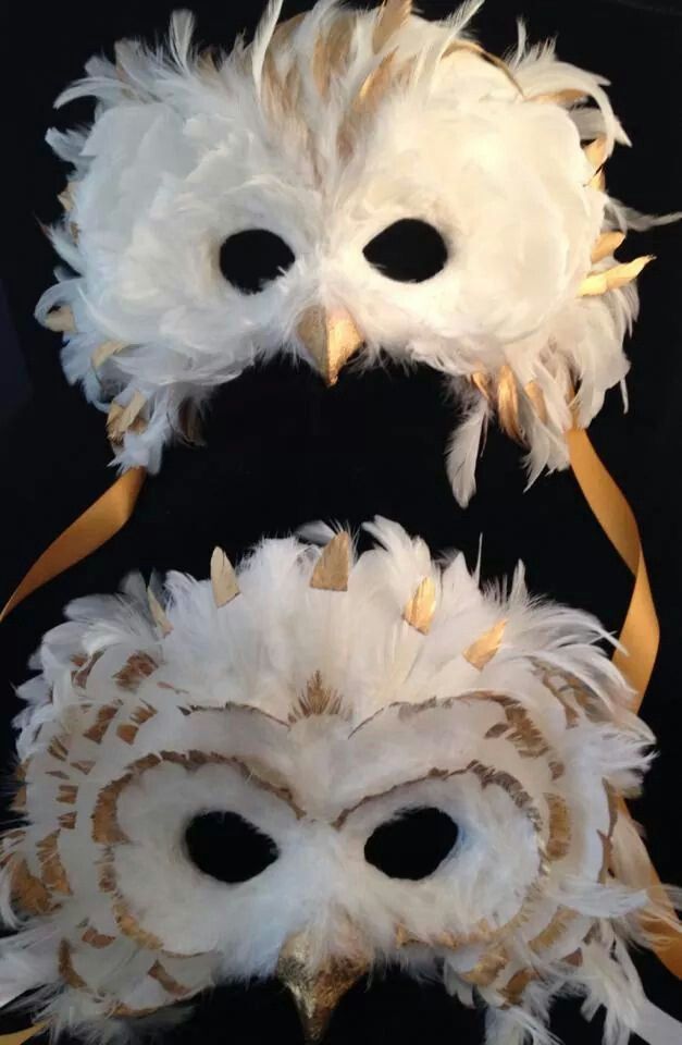 two white masks with feathers and gold trims on top of a black cloth background