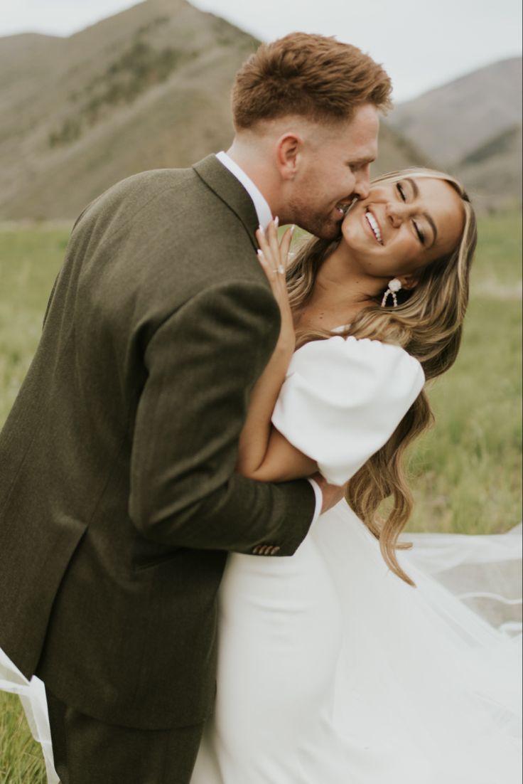 a bride and groom are kissing in the grass with mountains in the background