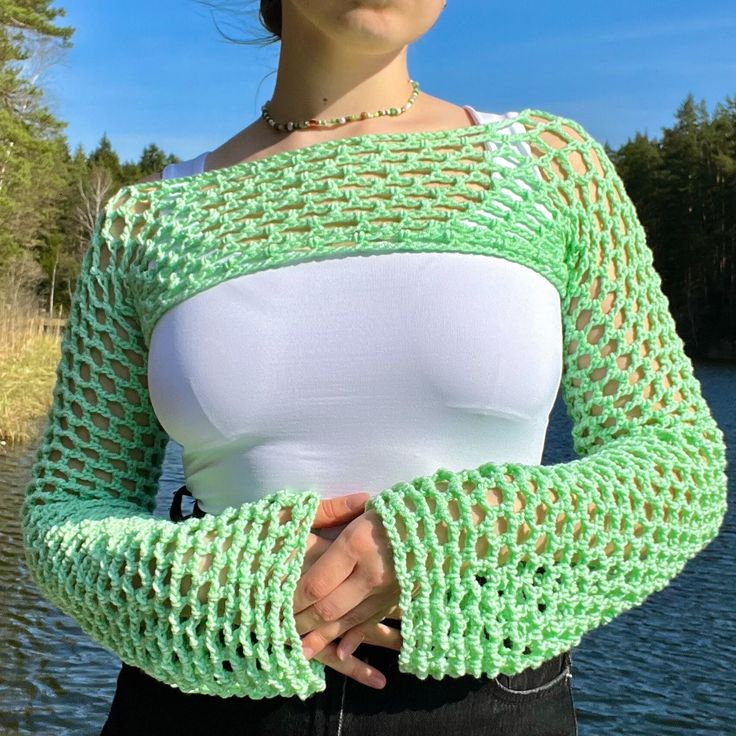 a woman standing next to a body of water wearing a green and white top with crocheted sleeves