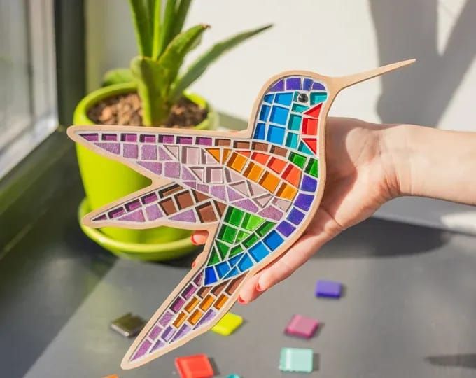 a hand holding a colorful bird shaped wooden puzzle next to a potted plant on a windowsill