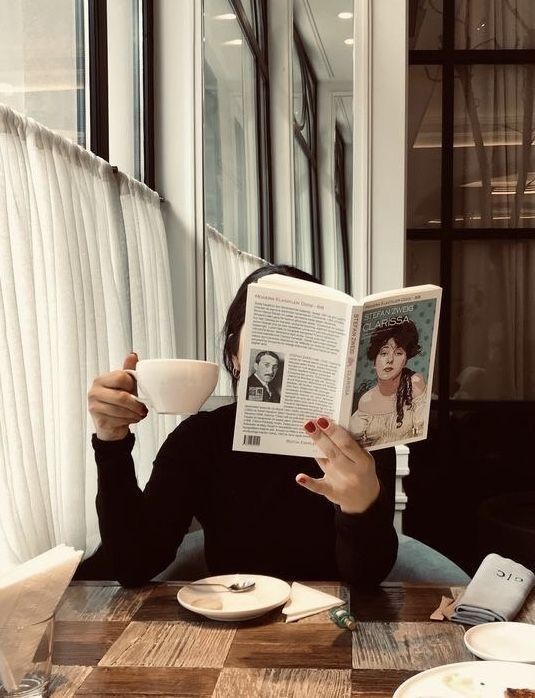 a woman sitting at a table reading a book and holding a coffee cup in front of her face