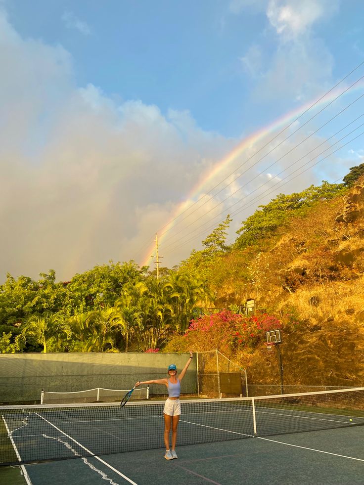 a woman standing on top of a tennis court holding a racquet under a rainbow