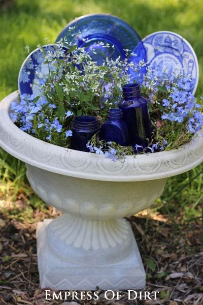 blue glass bottles and flowers in a white bowl on the ground with grass behind it