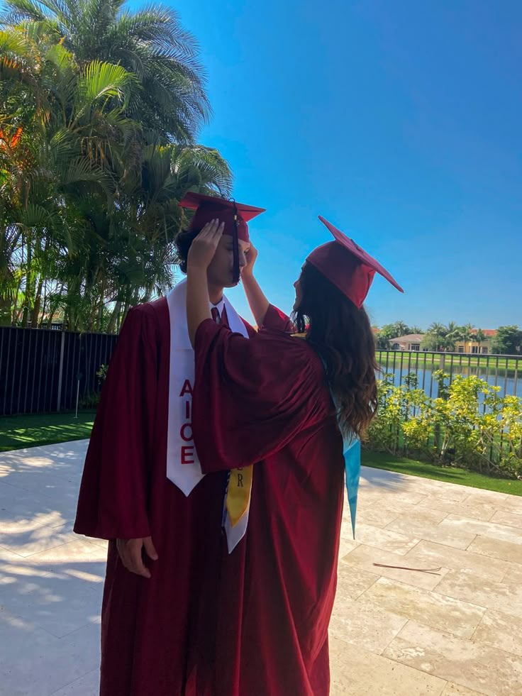 two people in graduation gowns and caps are looking at each other while one person adjusts the cap