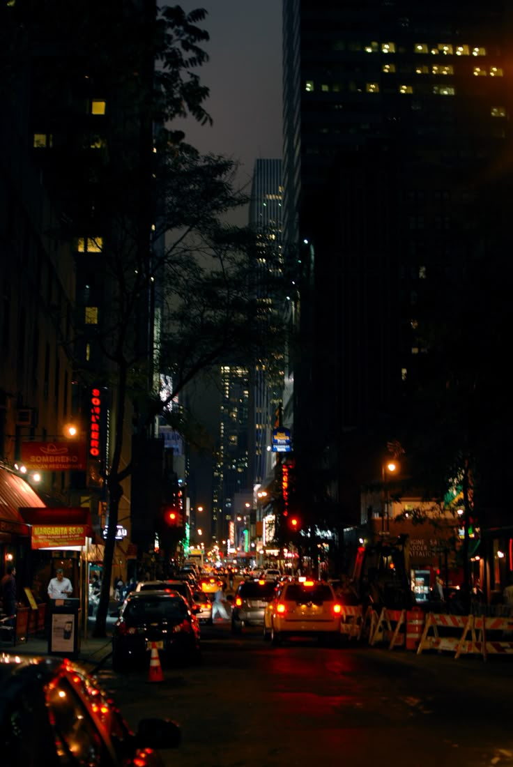 a city street at night with cars parked on the side and tall buildings in the background