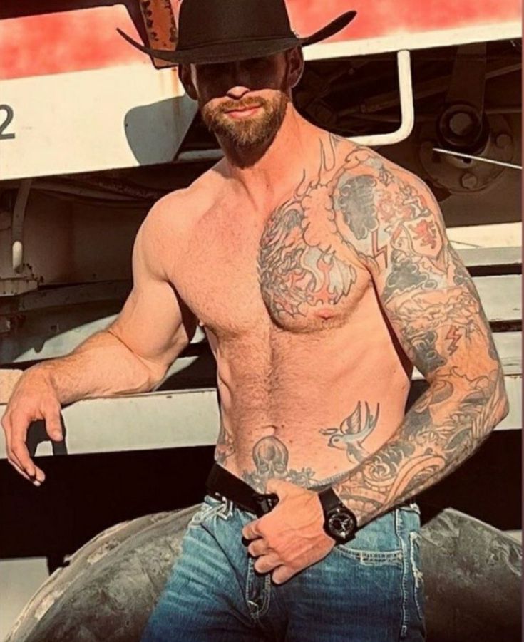 a shirtless man with tattoos and a cowboy hat sitting in front of a truck