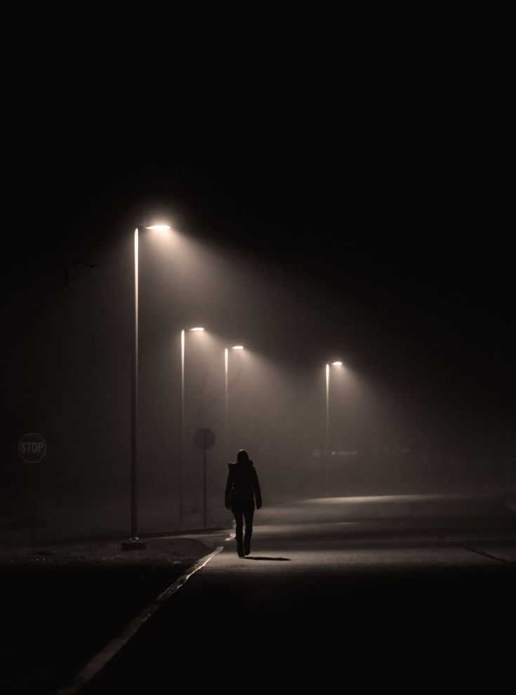 a person walking down a street at night in the fog with lights on either side