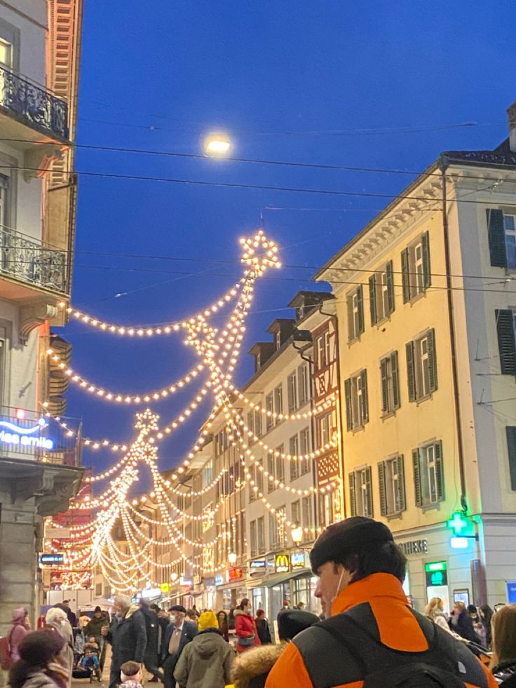 many people are walking down the street with christmas lights strung all over them and buildings in the background