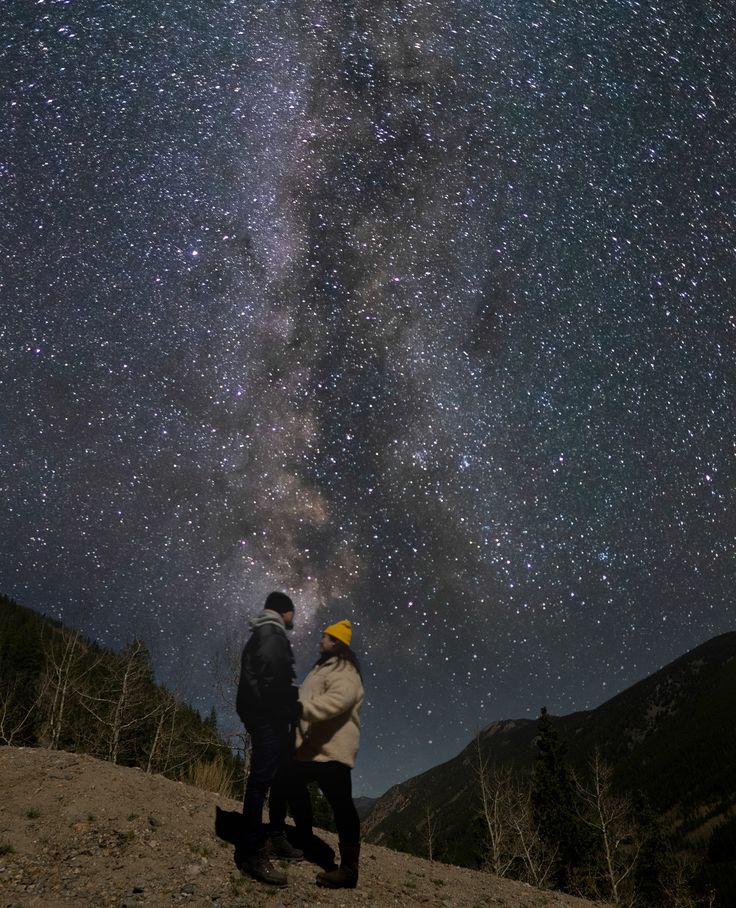 two people standing on top of a hill under the night sky with stars above them