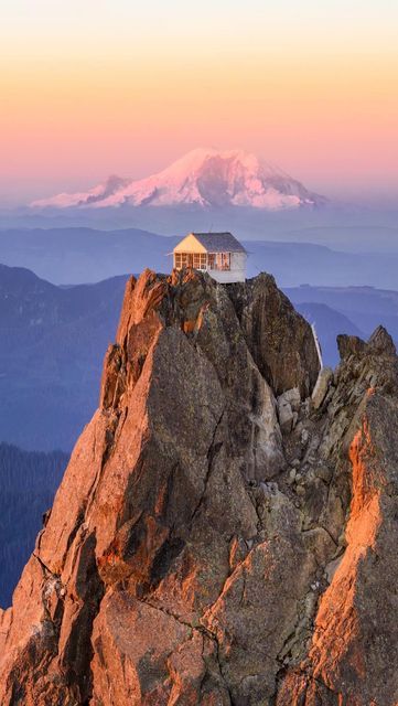 a house on top of a mountain with the sun setting in the sky behind it