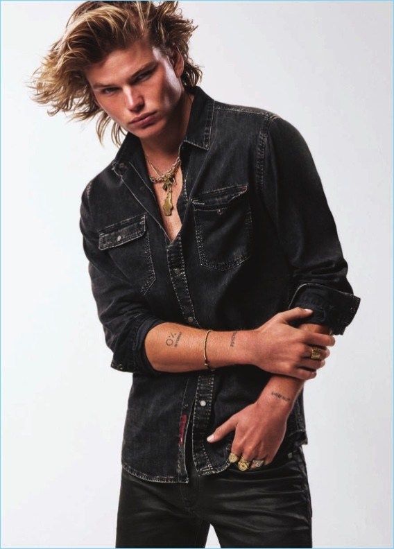 a young man with long hair wearing a denim shirt and black pants, standing in front of a white background