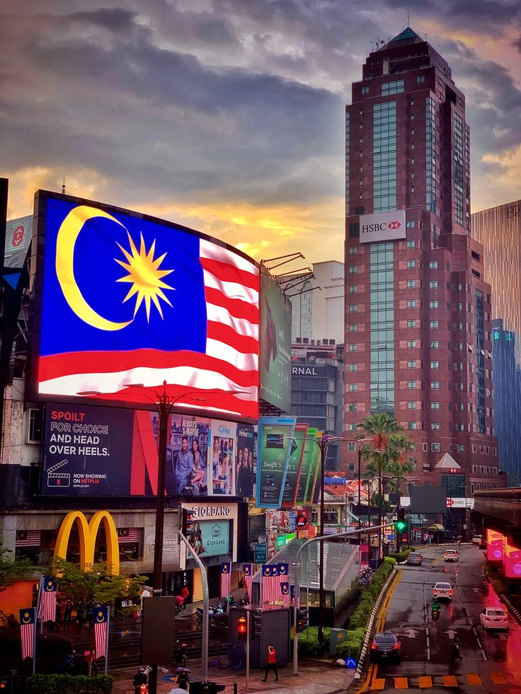 an american and malaysian flag in the middle of a city at sunset with tall buildings