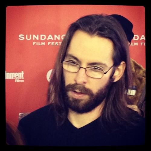 a man with long hair and glasses standing in front of a red wall at an event