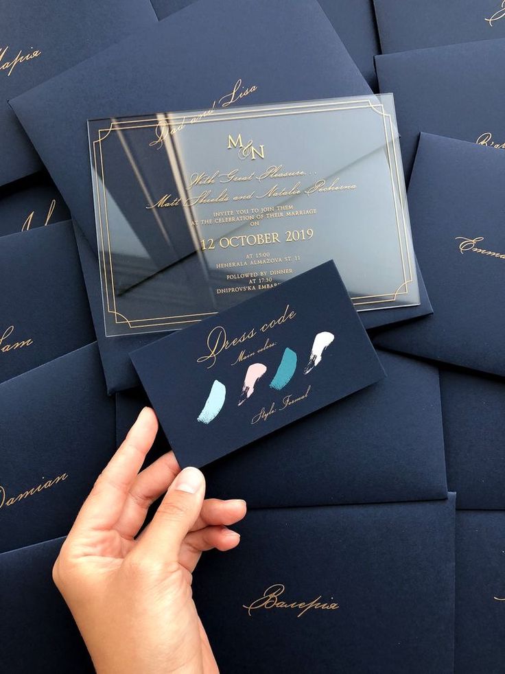 a person holding up a blue and gold wedding card in front of many black envelopes