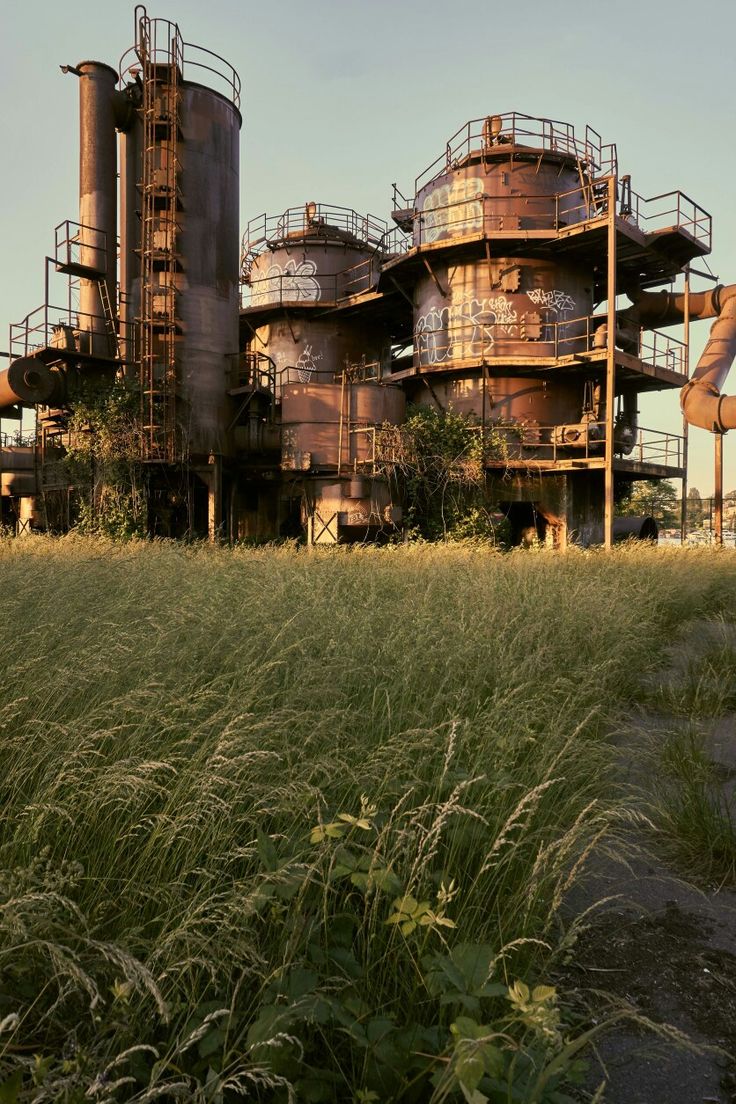 an old factory building with pipes and tanks in the foreground, surrounded by tall grass