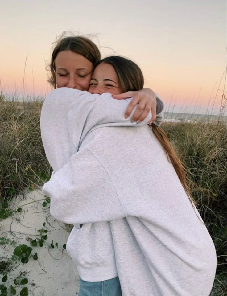 two women hugging each other on the beach at sunset or sunrise with sea oats in the background