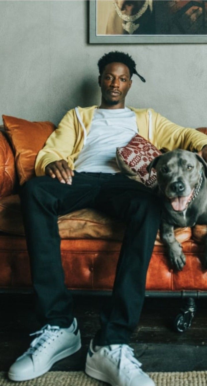 a man sitting on a couch next to a dog and wearing white sneakers with his tongue out