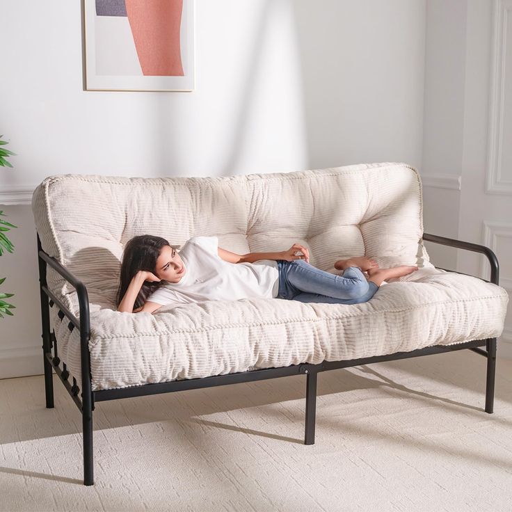 a woman laying on top of a white futon couch in a living room next to a potted plant