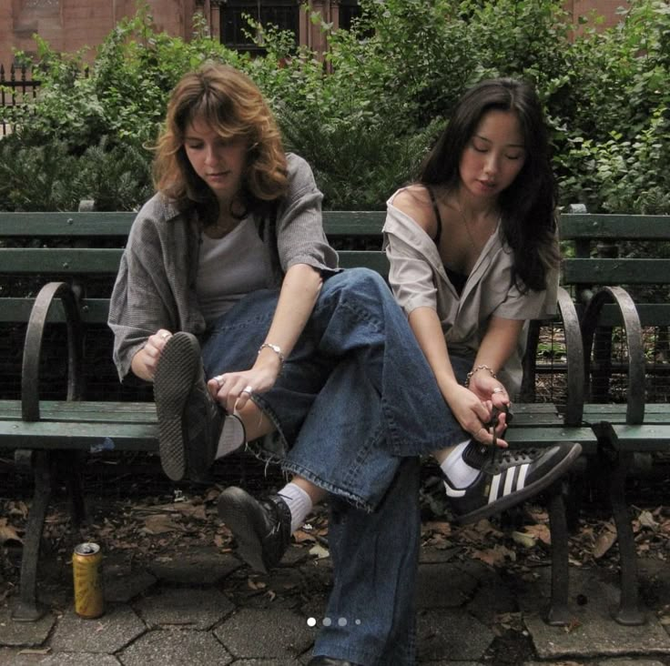 two young women sitting on a bench in front of some bushes and trees, one is holding her shoes