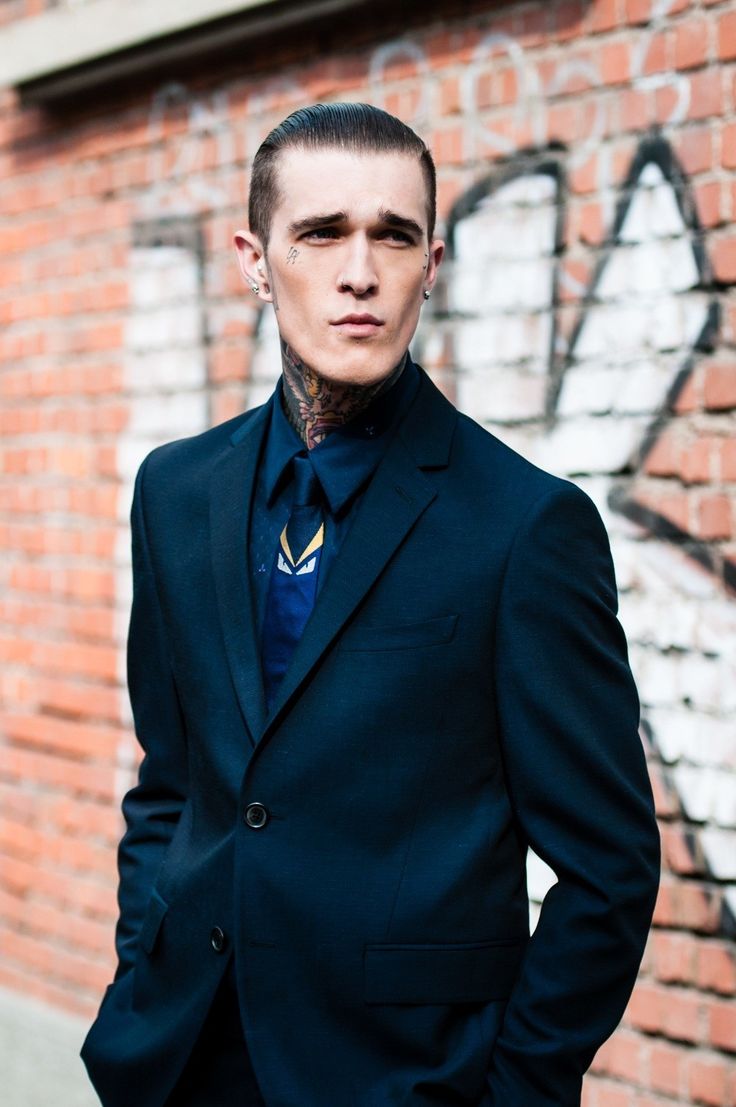 a man standing in front of a brick wall wearing a black suit and blue tie