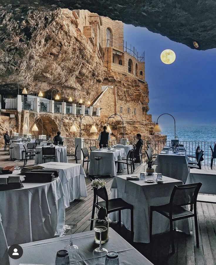 an outdoor dining area with tables and chairs overlooking the ocean at night, under a cliff