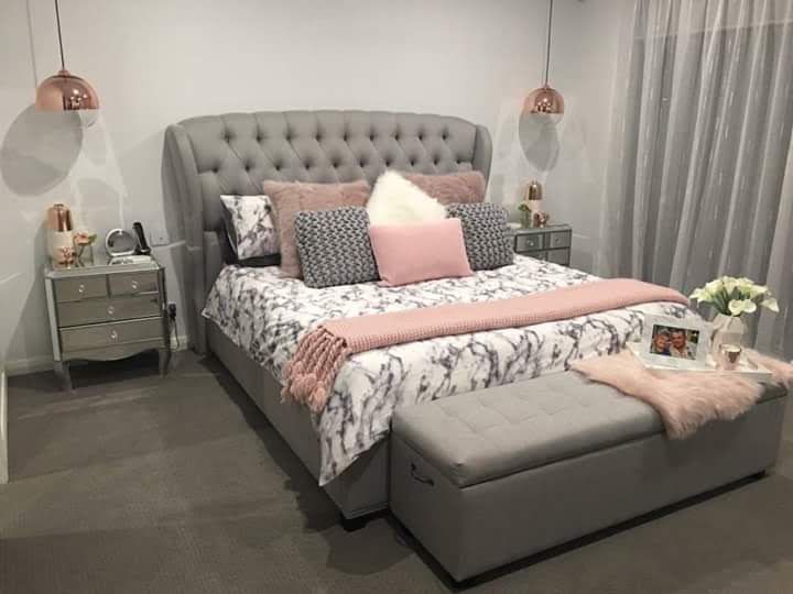 a bedroom with a gray bed, pink and white pillows, two nightstands and a grey ottoman