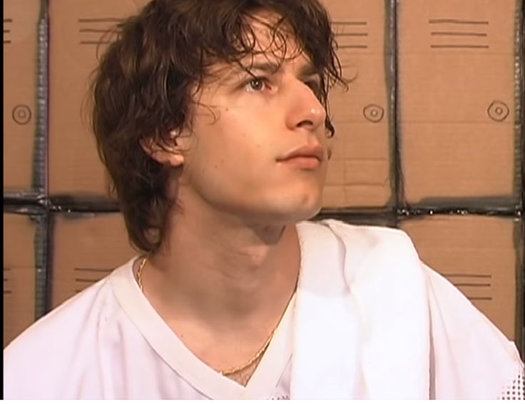 a young man with curly hair wearing a white shirt in front of stacks of boxes