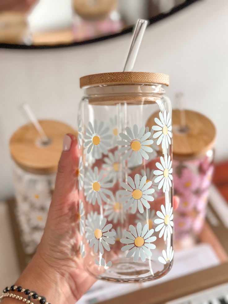 a person holding a mason jar with daisies painted on it and a straw in the lid