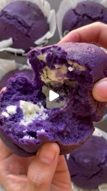 a person is holding up a purple doughnut with white stuff in it and there are other muffins behind them