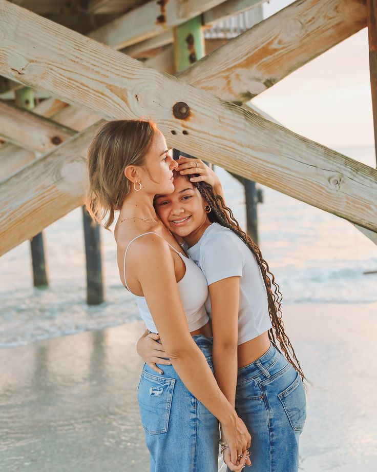 two young women hugging each other under a wooden structure on the beach with water in the background