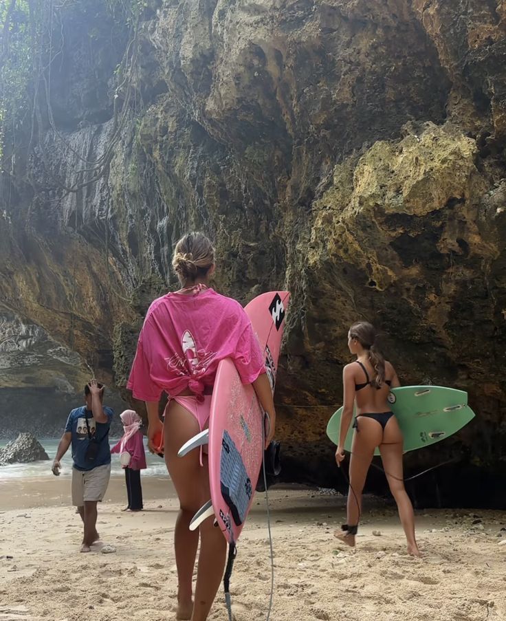 two women in bikinis are carrying surfboards on the beach while others walk by