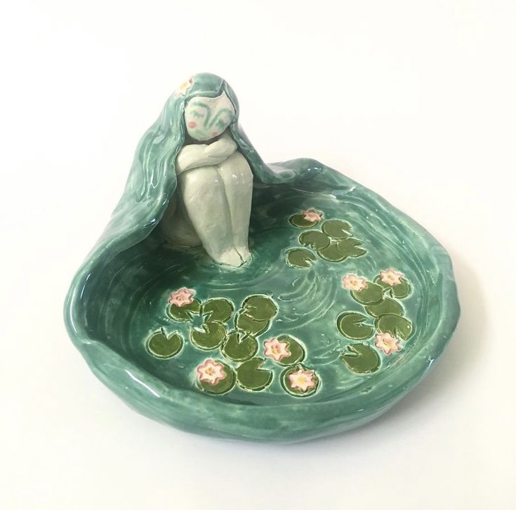 a ceramic figurine sitting on top of a green bowl filled with water lilies