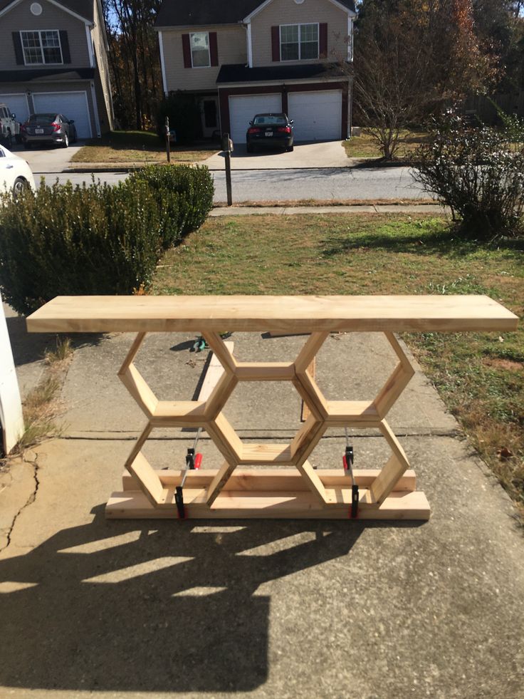 a wooden table sitting on top of a sidewalk next to a yard with cars parked in the background