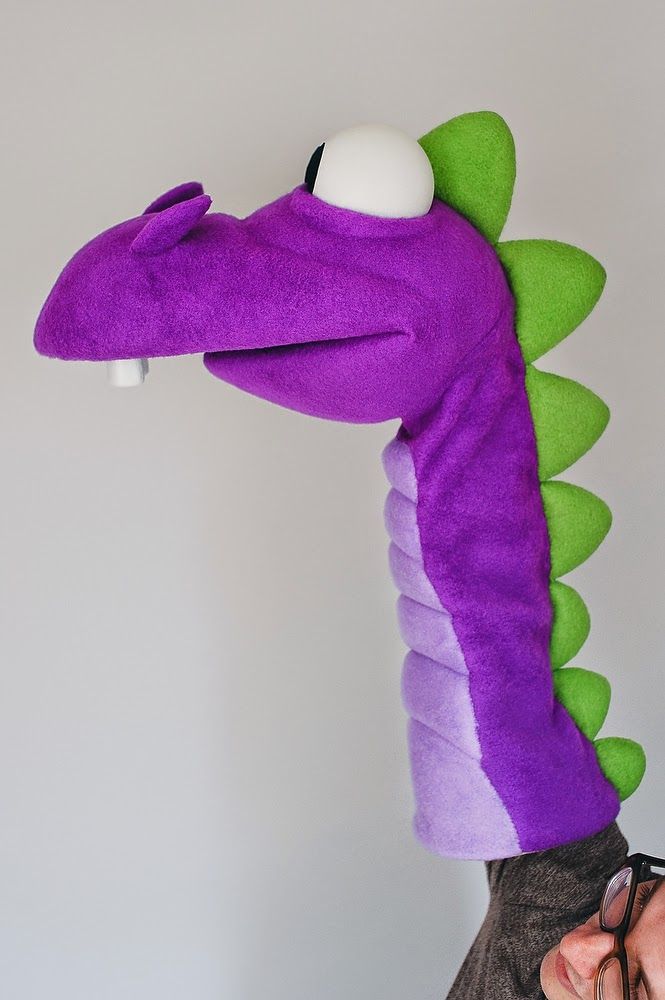 a purple and green stuffed animal with eye glasses on it's head next to a white wall